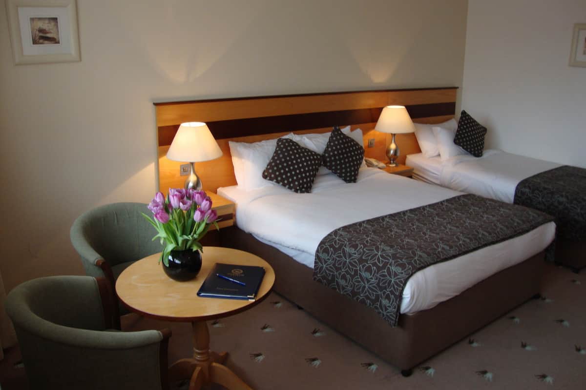 A hotel room with two beds, a chair, a small table with a vase of flowers, and a book.