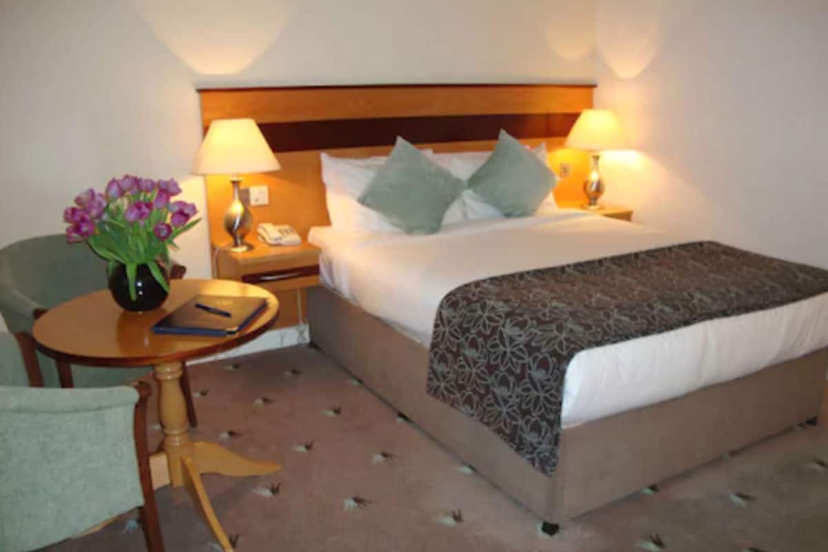 A hotel room with a bed, bedside lamps, a small table with a vase of flowers, a chair, and a carpet with a pattern.