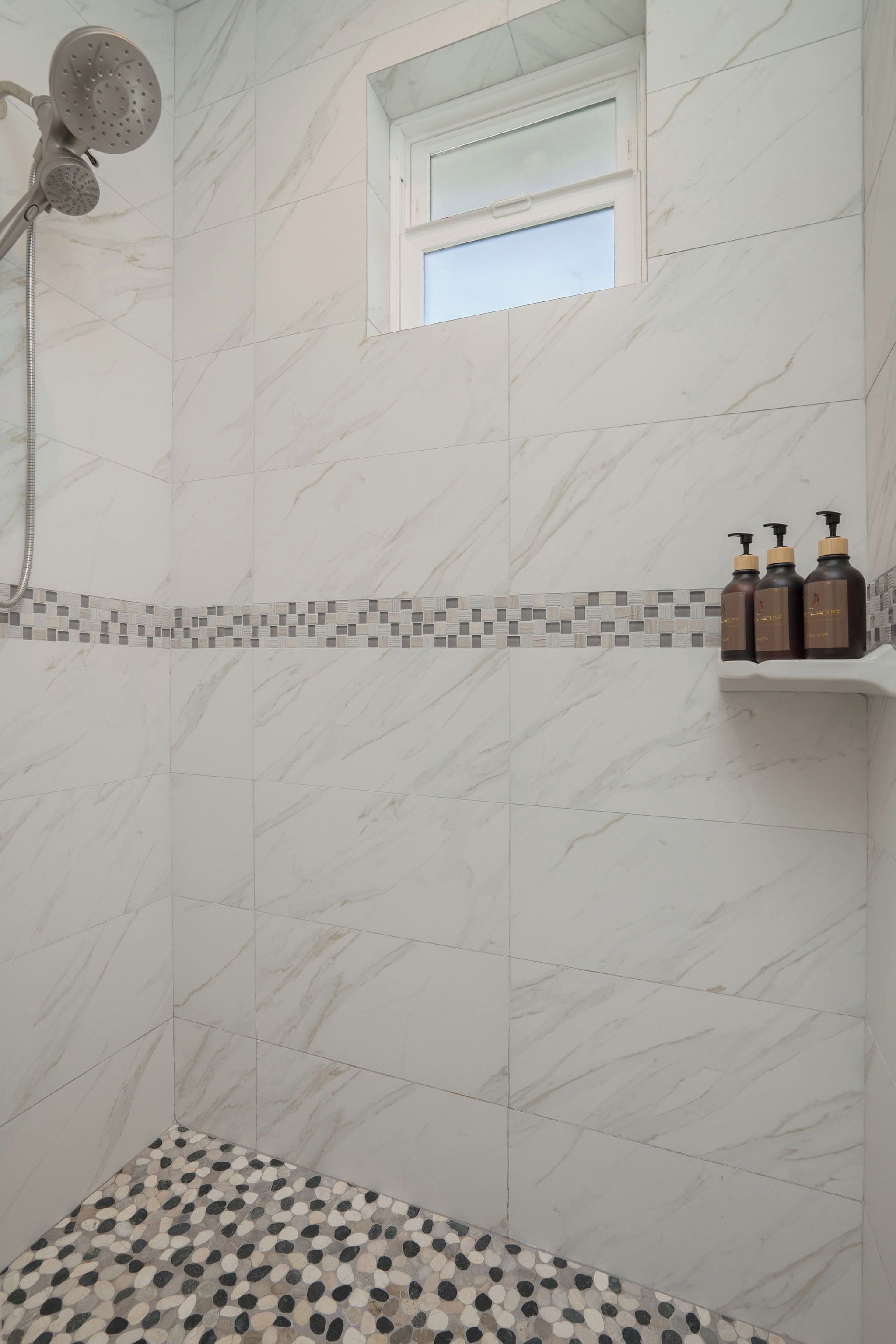 A bathroom with white marble tiles, a showerhead, a small window, and toiletries on a shelf.