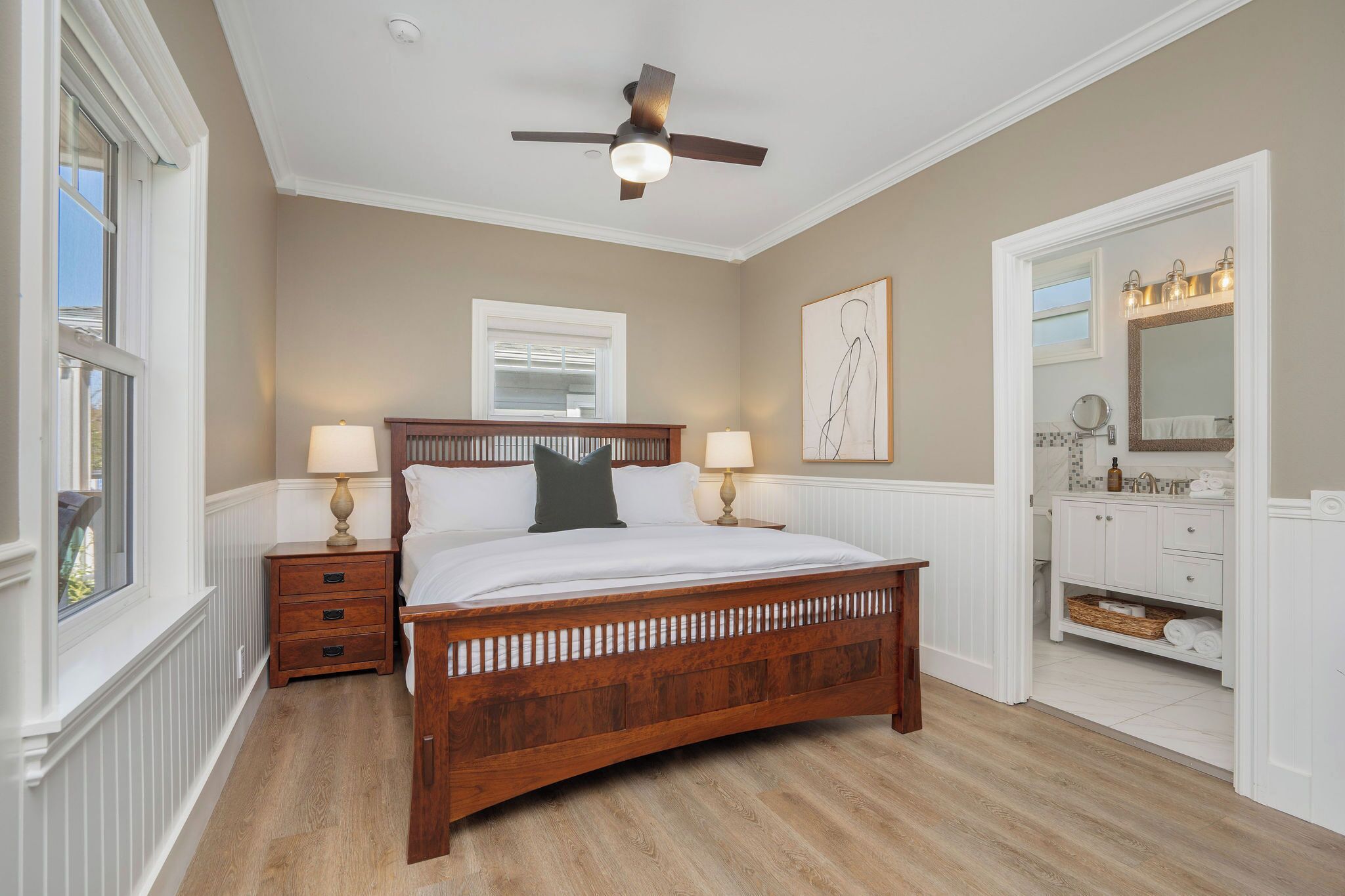 A bedroom with a wooden bed, two bedside tables with lamps, a ceiling fan, a window, a bathroom with a vanity and mirror.