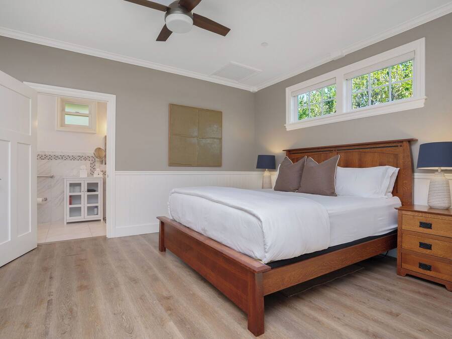 Un dormitorio con cama de madera, una mesita de noche, ventilador de techo, ventana con vistas y puerta que da a otra habitación.