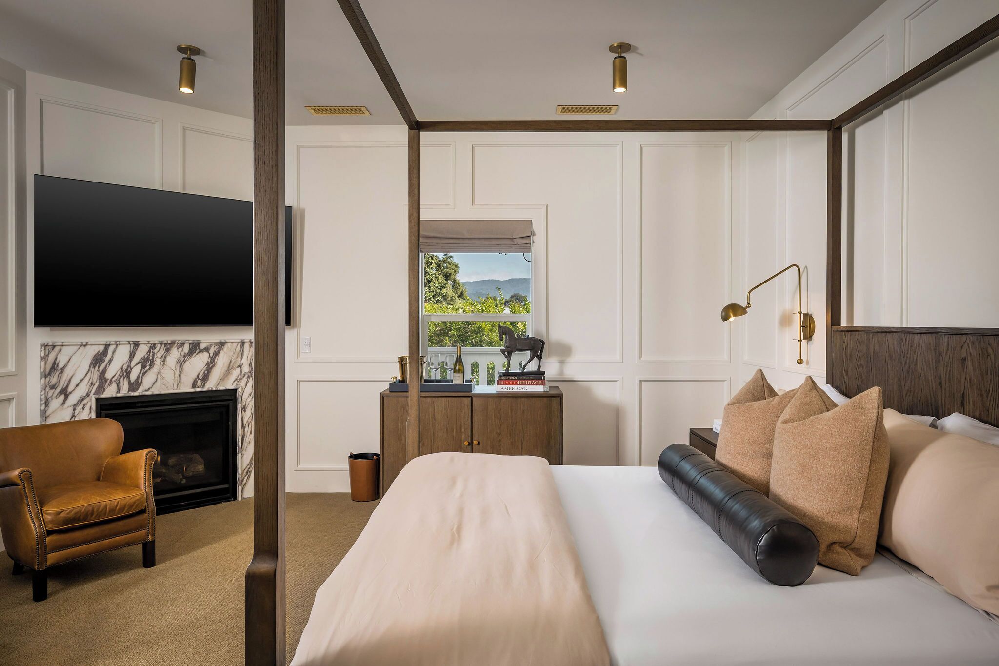 Habitación de hotel con una cama grande, televisión montada en la pared, chimenea, mesita de noche de madera y vistas al exterior.