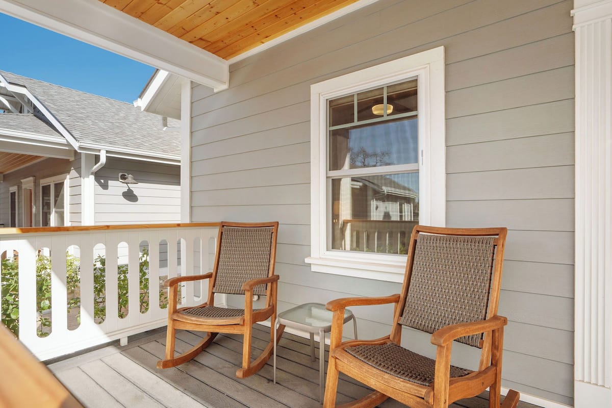 A porch with two rocking chairs and a small table, wooden railing, and a window with a view of the sky.