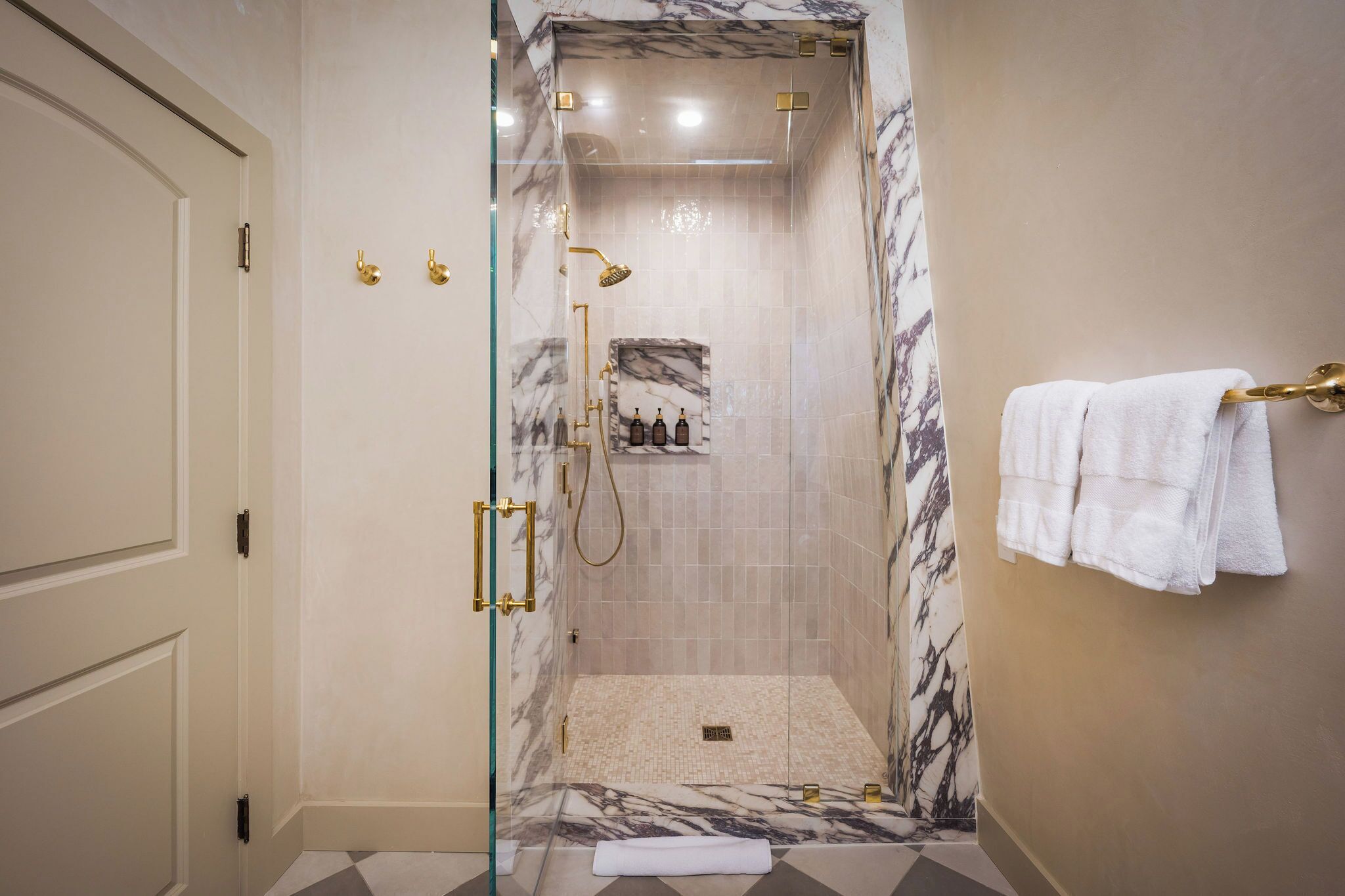 Ducha con cerramiento de vidrio, paredes de mármol, un perchero con toallas y una puerta con pomo de latón.