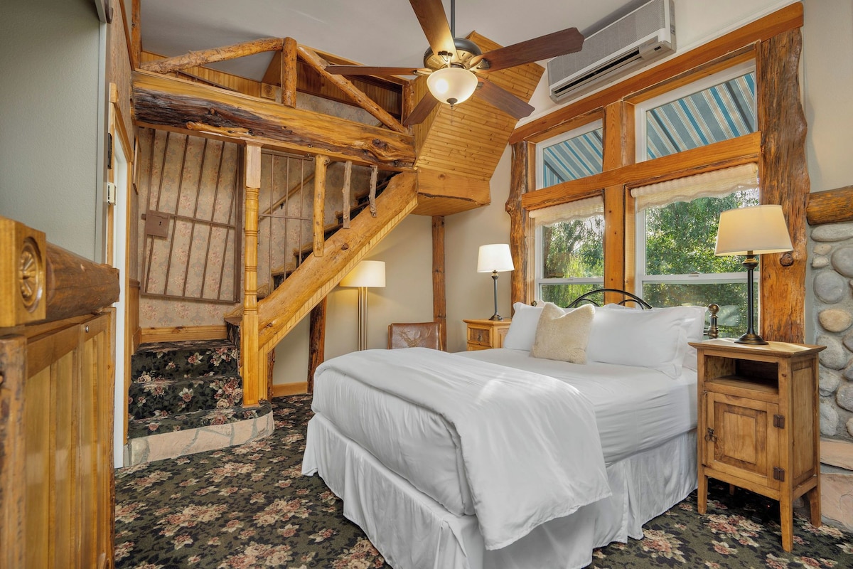 A bedroom with a wooden staircase, a bed with white bedding, two bedside lamps, and a stone wall.