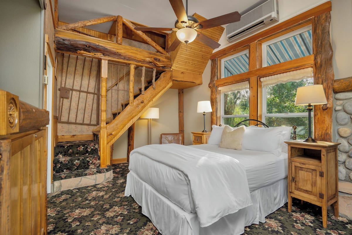 A bedroom with a wooden staircase, a bed with white bedding, two bedside lamps, and a stone wall.