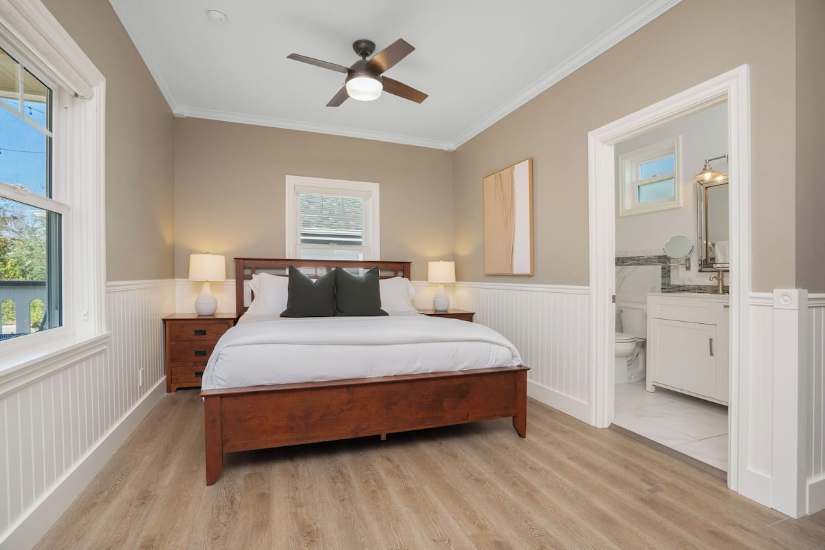 A bedroom with a wooden bed, two bedside tables with lamps, a ceiling fan, a large window, and a view of the outside.