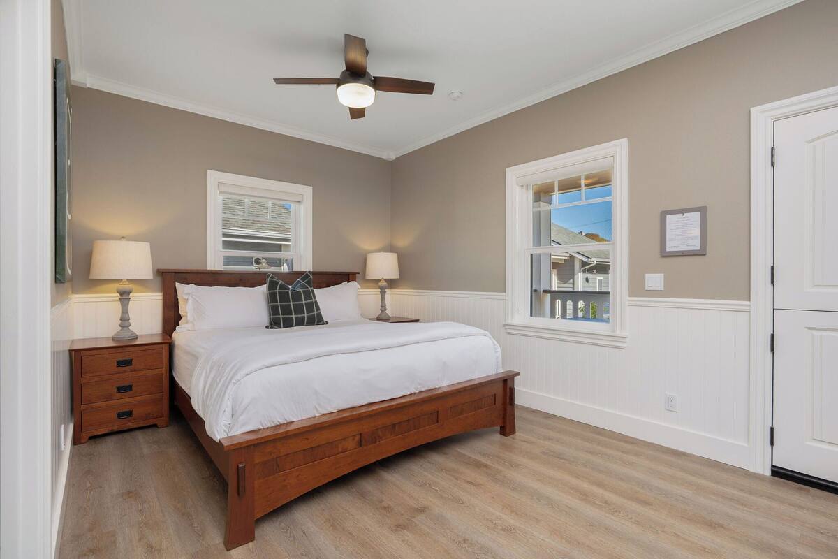 A bedroom with a wooden bed, two bedside tables with lamps, a ceiling fan, and a window with a view of the outside.
