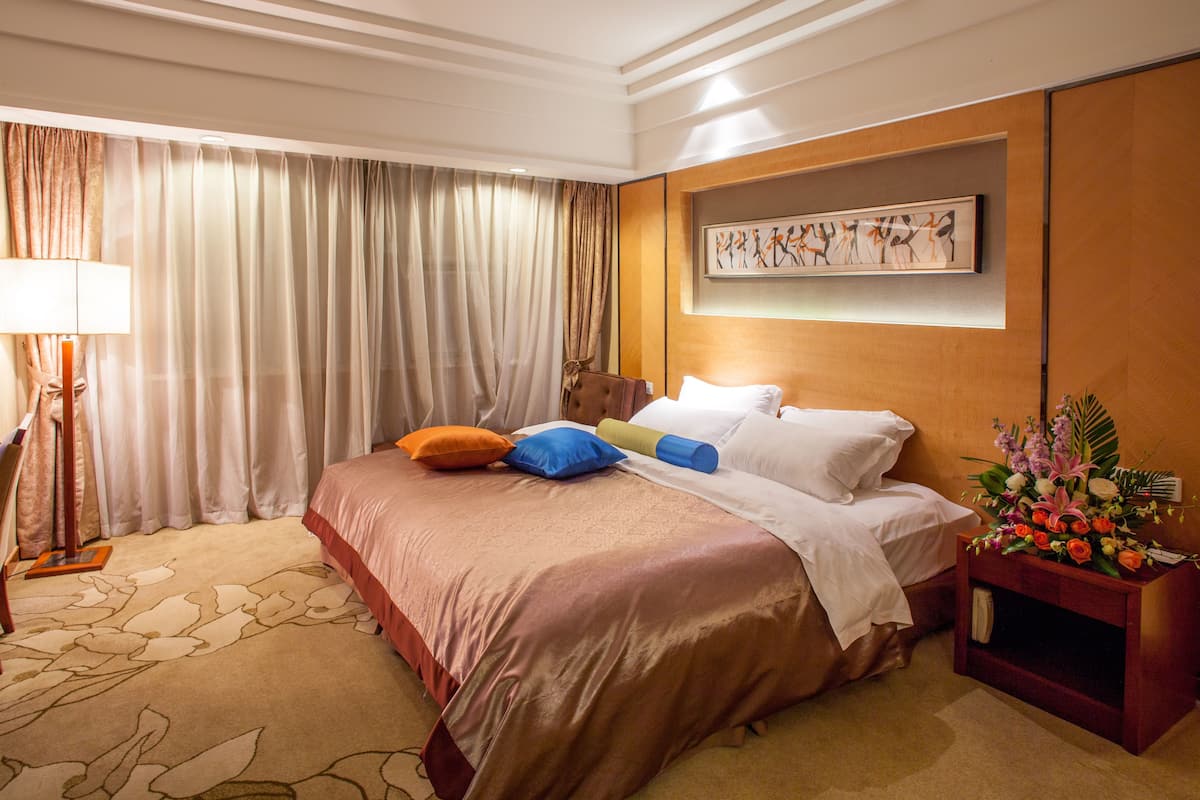 Quarto de hotel com uma cama grande, cabeceira de madeira, uma obra de arte emoldurada acima da cama, mesa de cabeceira com um vaso de flores e uma janela com cortinas.