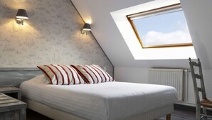 Un dormitorio con techo inclinado, tragaluz, cama con almohadas a rayas, dos lámparas fijadas a la pared y un radiador.