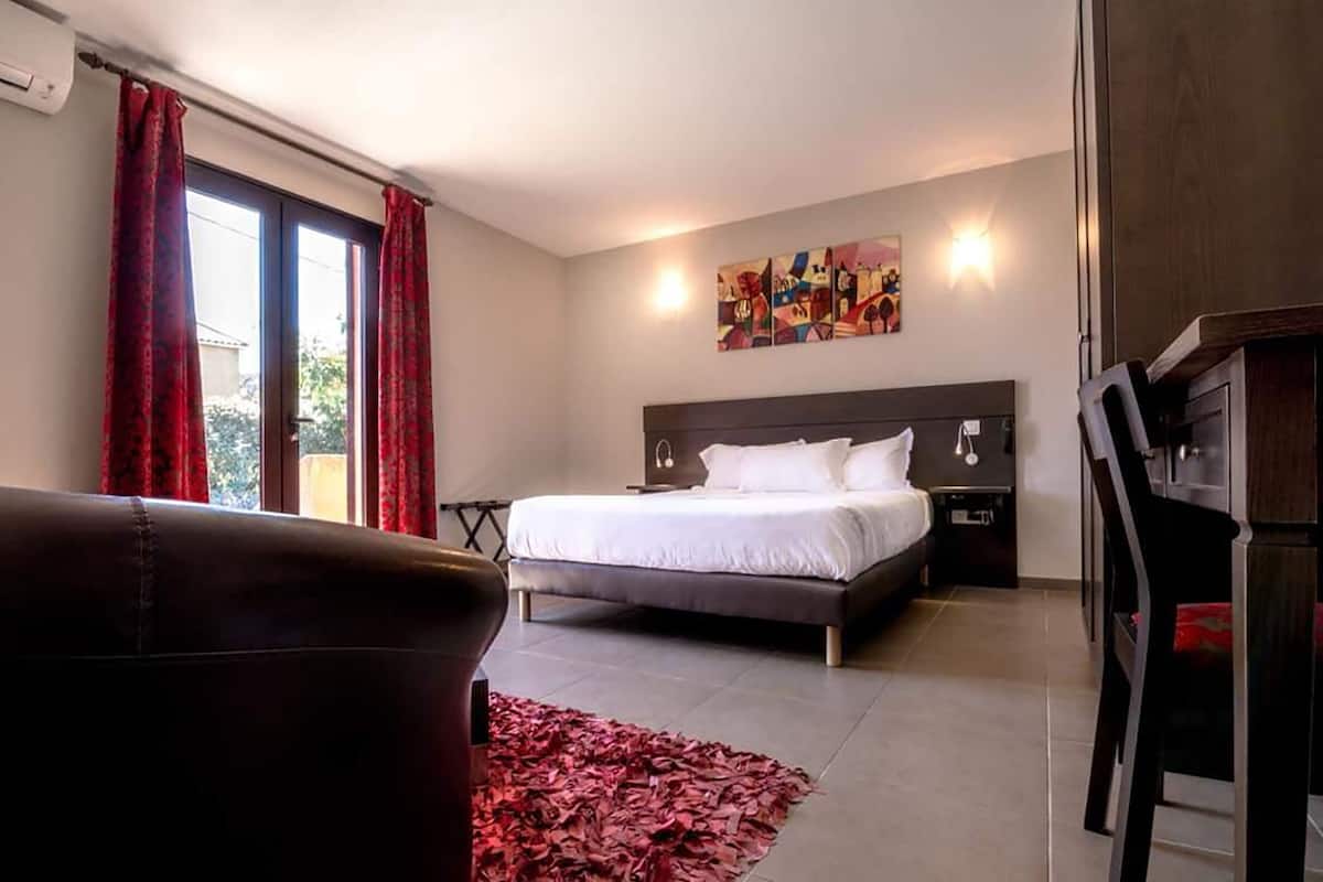 Um quarto com uma cama, mesa de cabeceira, escrivaninha, cadeira e uma janela grande com cortinas.