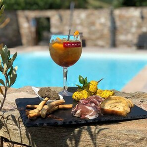 Una copa de vino L.N. Mattei, una pizarra de pizarra negra con varios quesos, galletas y embutidos, y una piscina al fondo.