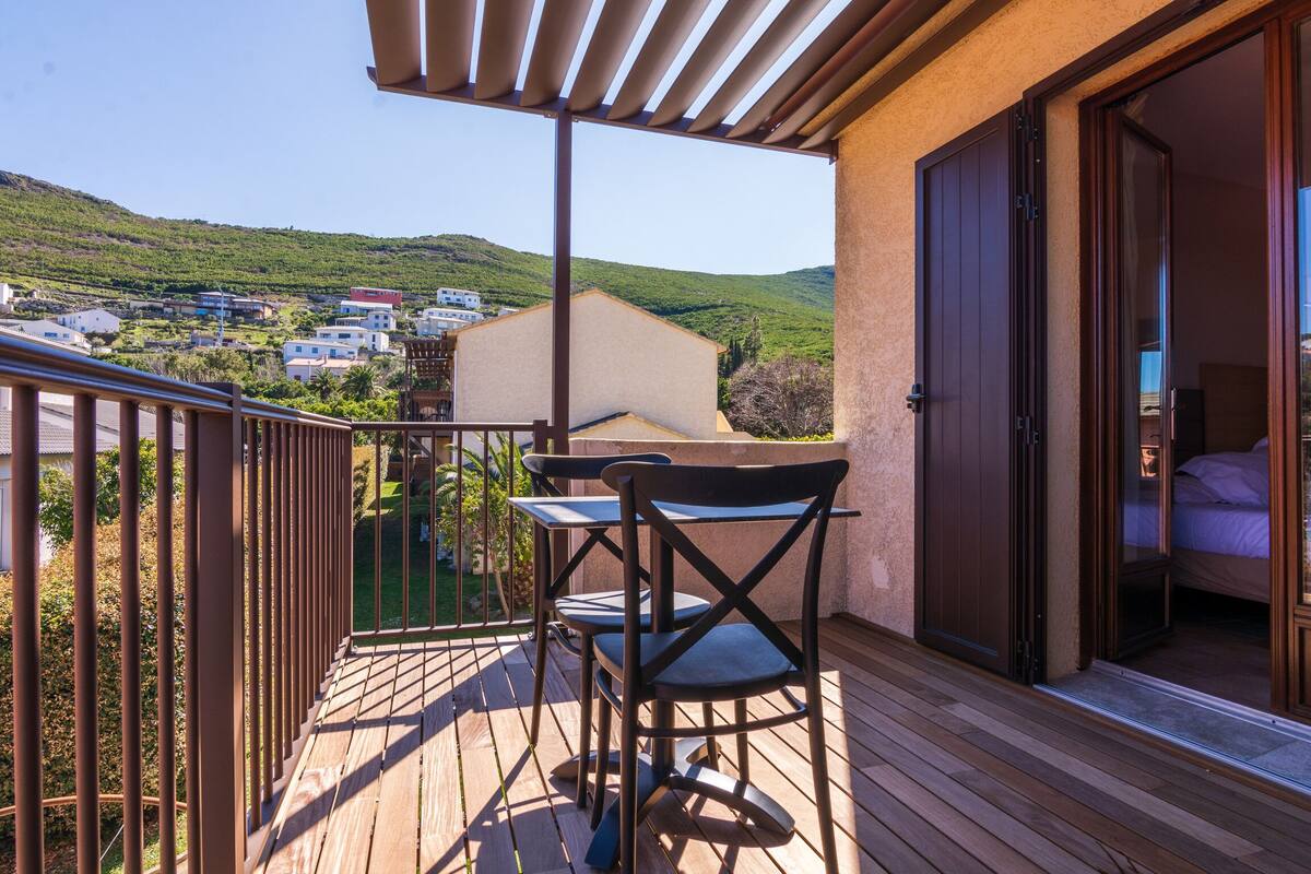 Uma varanda com mesa e cadeiras, uma porta de correr de vidro que dá para um quarto e vista para uma encosta com edifícios.