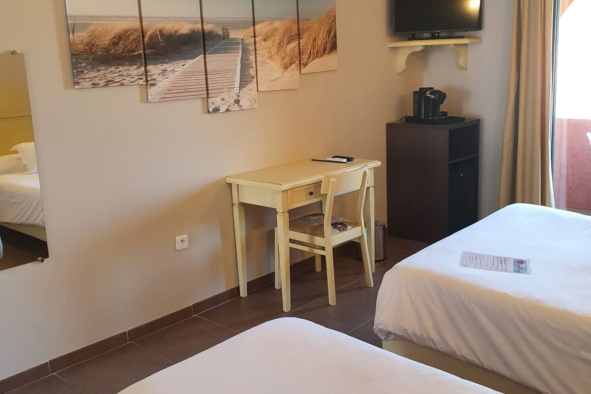 Quarto de hotel com duas camas, uma escrivaninha, uma cadeira, uma televisão e ar-condicionado fixado na parede.