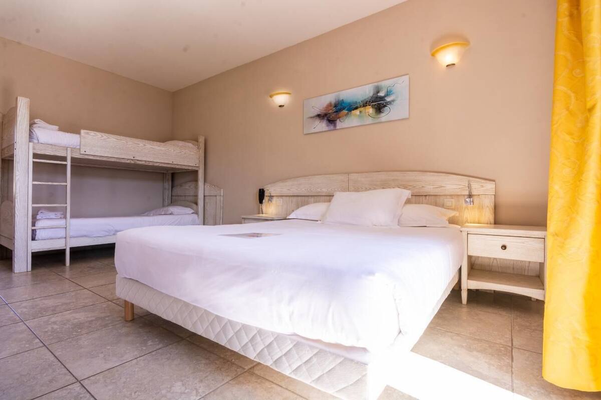 Quarto com uma cama grande, uma cama menor, uma beliche e uma mesinha de cabeceira com abajur.