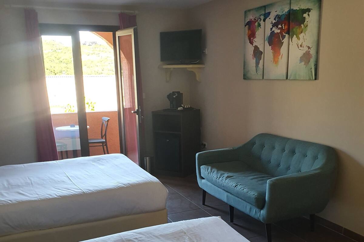Quarto de hotel com duas camas, um sofá verde-água, uma TV sobre um suporte e um mapa-múndi na parede.
