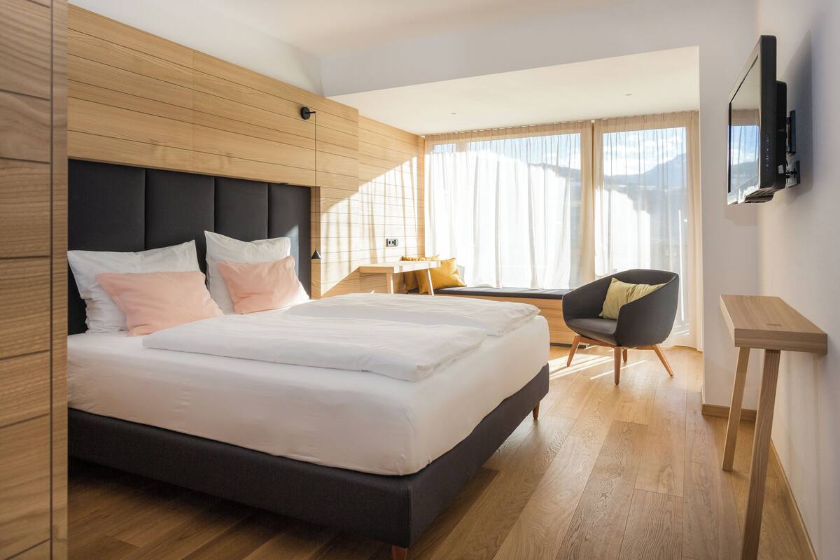 A modern bedroom with a large bed, a wooden headboard, a small table, a chair, and a TV mounted on the wall.
