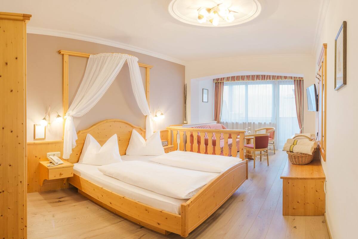 A wooden four-poster bed with white bedding, a bedside table with a lamp, a wooden nightstand, a wicker basket, a wooden bench with a cushion, and a television on the wall.