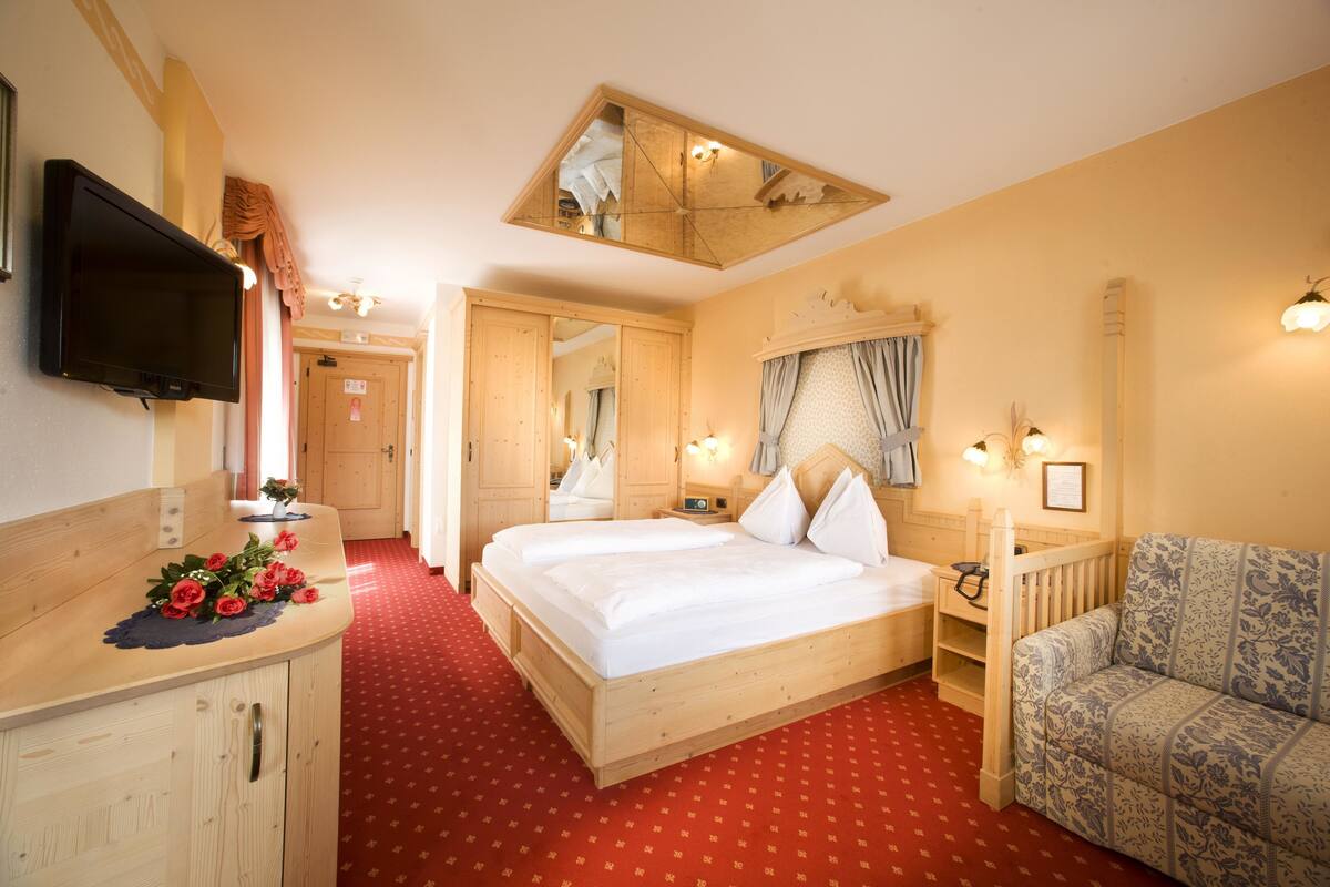 A hotel room with a wooden bed, a TV mounted on the wall, a red carpet, a wooden dresser, and a floral-patterned armchair.