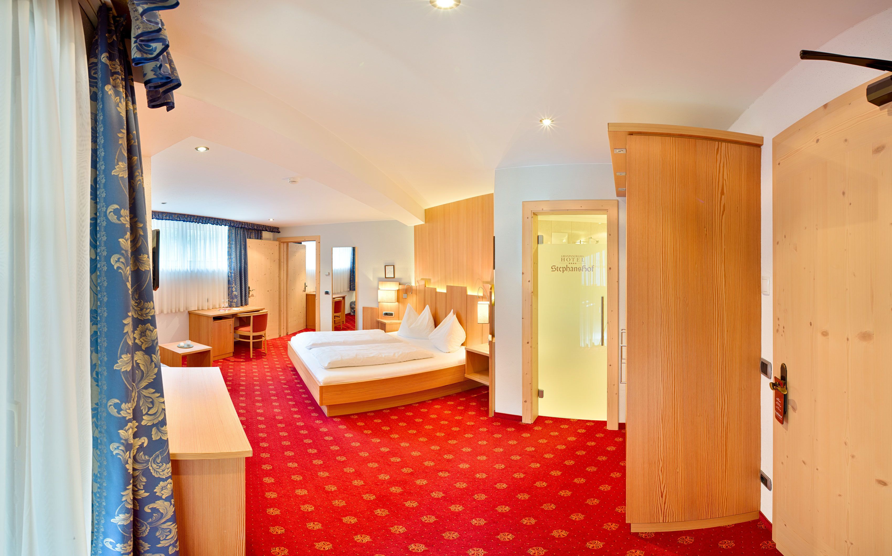 A hotel room with a bed, desk, chair, and a red carpeted floor.