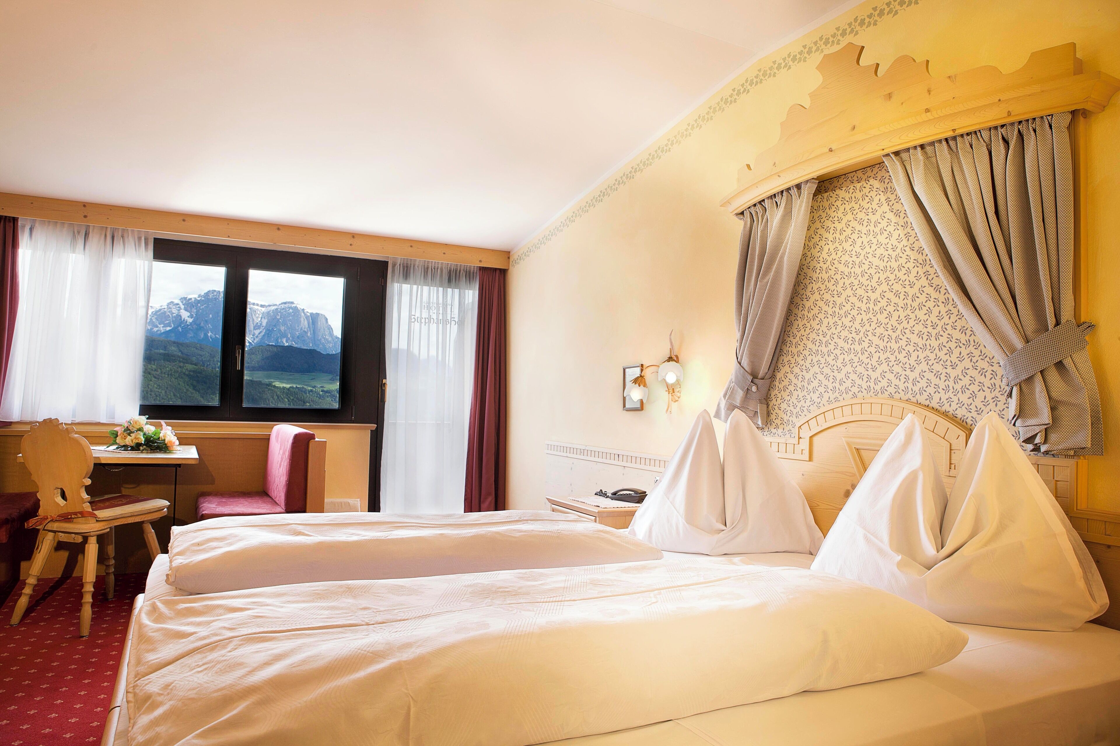 A hotel room with a large bed, a desk, and a chair. There is a window with a mountain view.