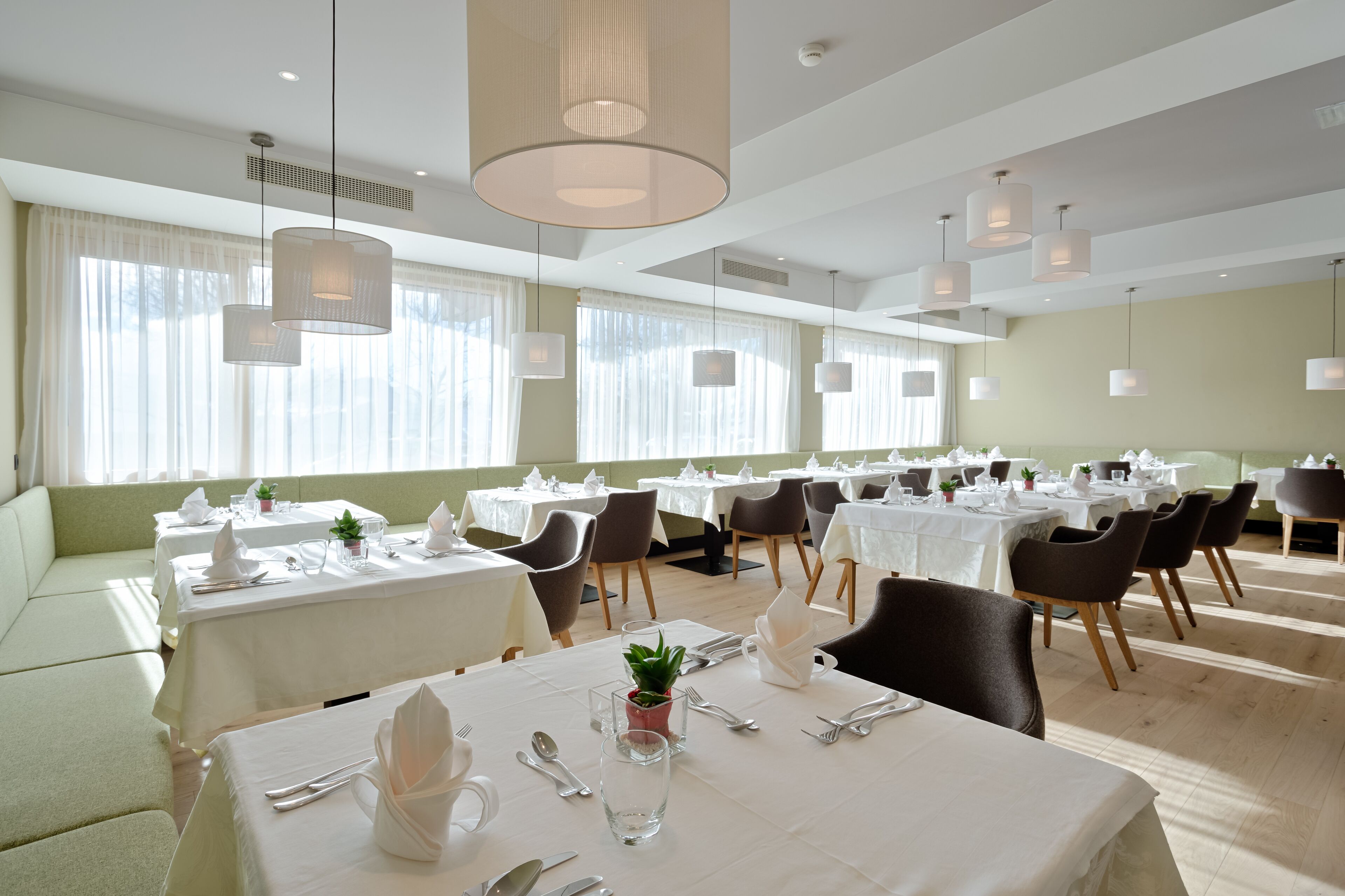 A restaurant with multiple tables set for dining, featuring white tablecloths, neatly arranged cutlery, and hanging pendant lights.