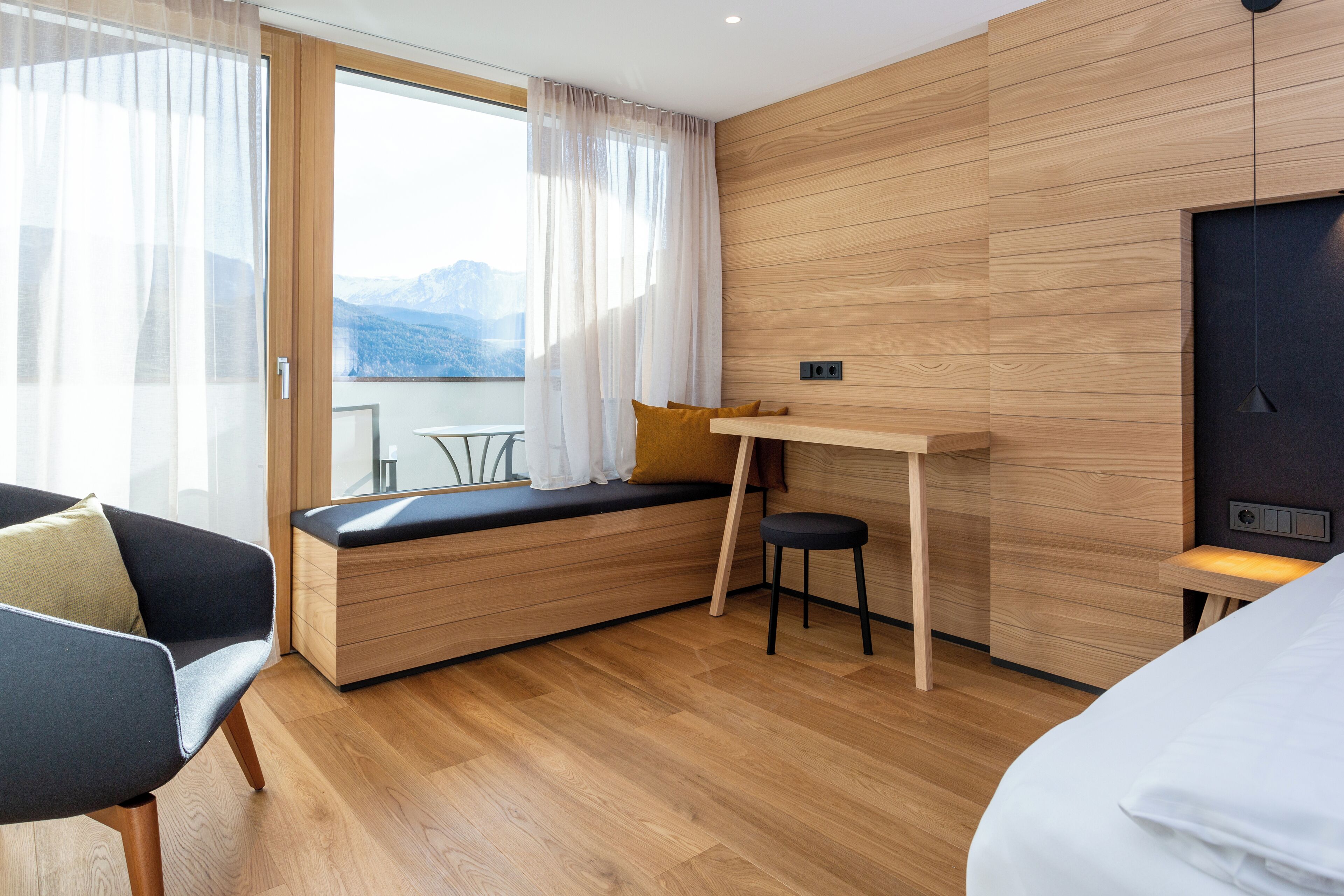 Ein modernes Zimmer mit Holzboden, einem Bett, einem Schreibtisch mit Stuhl, einem Fenster mit Bergblick und einer Couch mit Kissen.