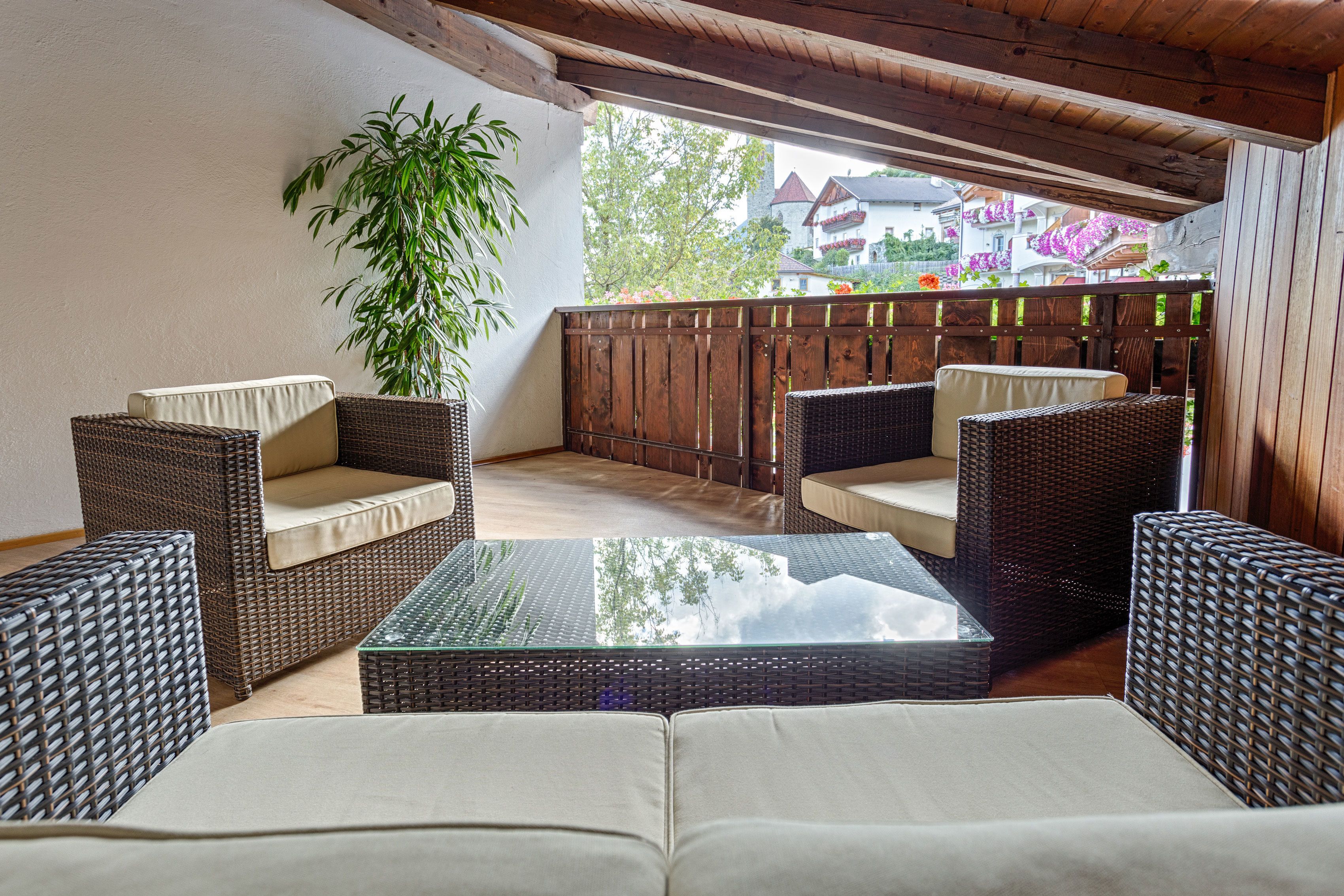 A covered outdoor seating area with wicker furniture and a glass table, overlooking a balcony with a wooden railing and a view of buildings and trees.