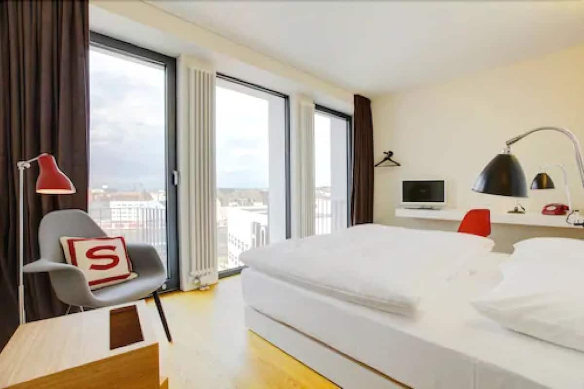 Hotellihuone, jossa on sänky, työpöytä, tuoli, televisio ja suuret ikkunat, joista avautuu näkymä kaupunkiin.