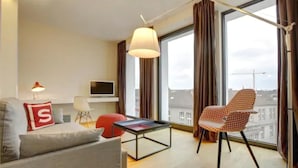 A modern living room with a sofa, a red cushion, a table, a chair, a TV, and large windows with curtains. - Smartcity Designhotel (Hannover)