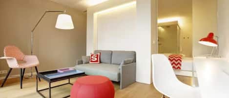 A modern living room with a grey sofa, a red ottoman, a white dining table, and a red lamp.