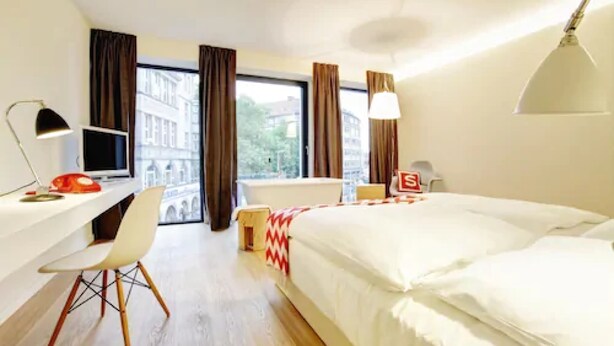A modern hotel room with a bed, desk, chair, TV, and a view of buildings through large windows. - Smartcity Designhotel (Hannover)