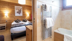 A wooden-paneled room with a bed, a nightstand, a wall-mounted heater, and a bathroom with a bathtub and a towel rack. - La Ferme du Lac (Thyez)