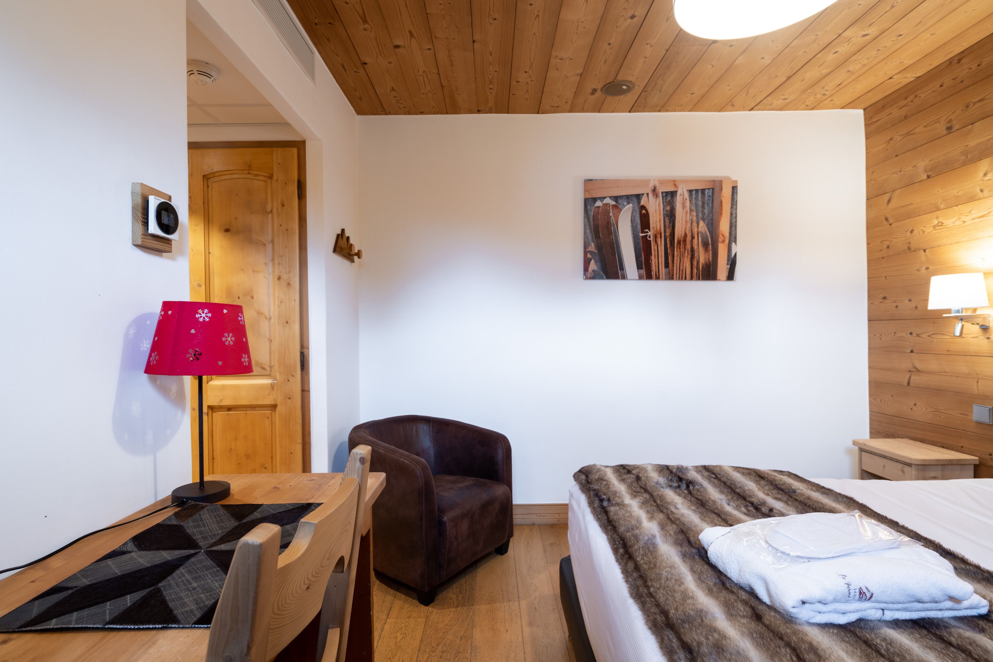 Una habitación acogedora con techo de madera, una cama con manta de aspecto aterciopelado, un sillón de cuero marrón, una mesa de madera y una lámpara roja.