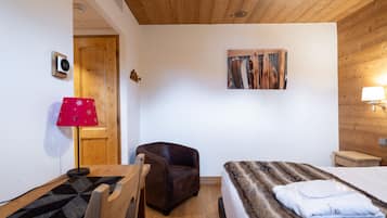 Una habitación acogedora con techo de madera, una cama con manta de aspecto aterciopelado, un sillón de cuero marrón, una mesa de madera y una lámpara roja.