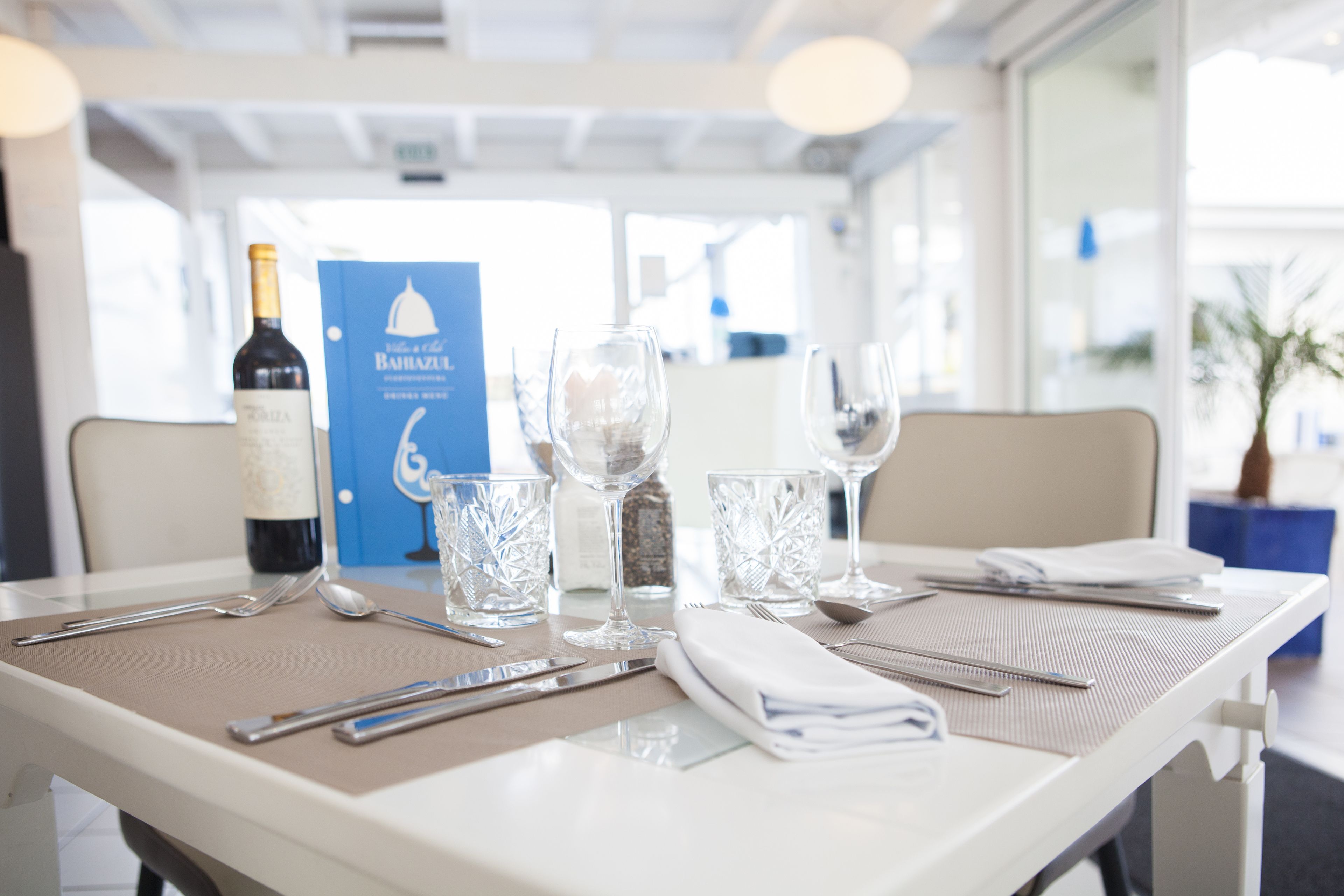 A table set with wine glasses, cutlery, and a bottle of wine, with a placemat and a blue informational board in the background.