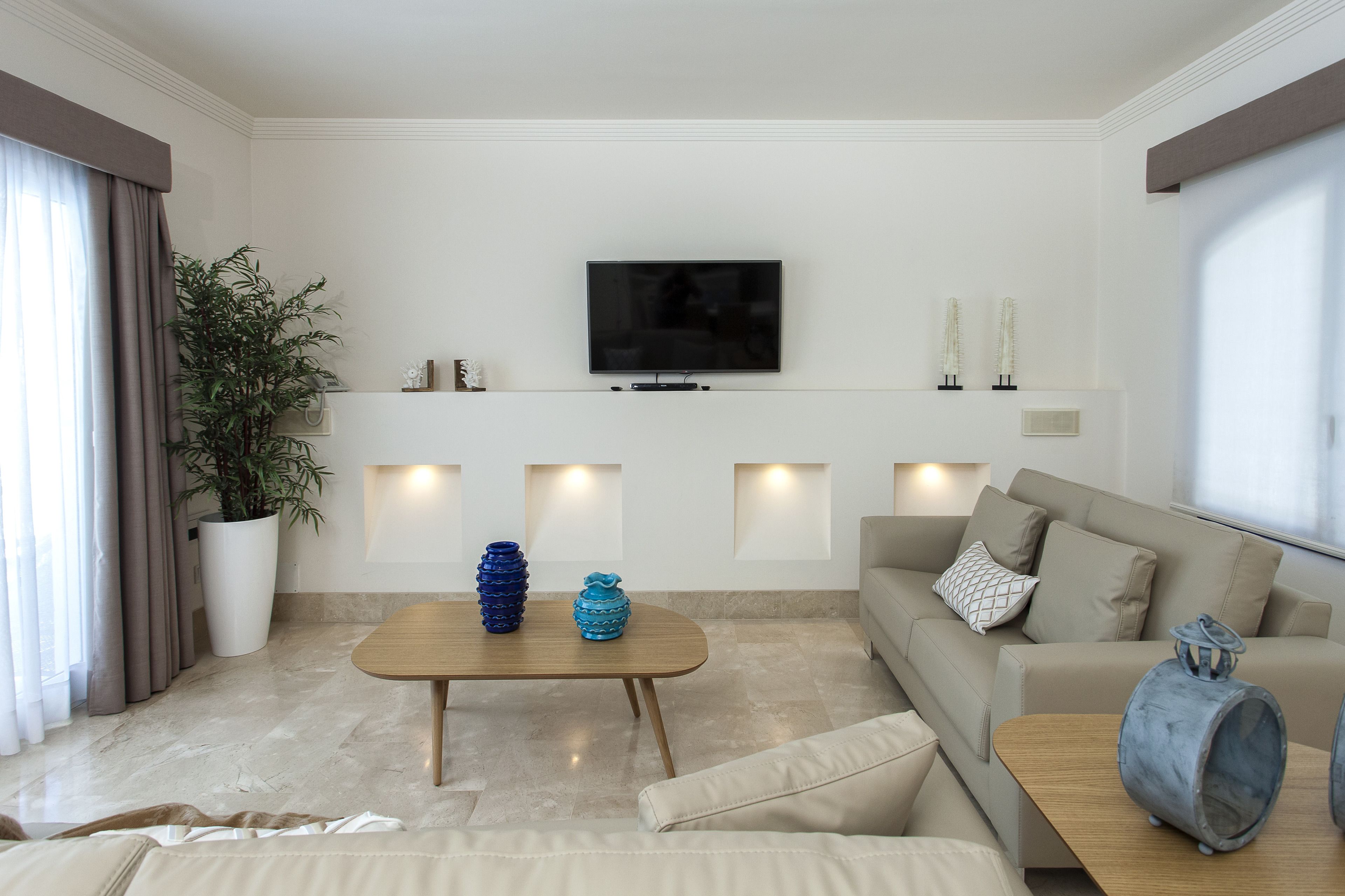 A modern living room with a flat-screen TV, a large potted plant, a wooden coffee table, and a beige sofa.
