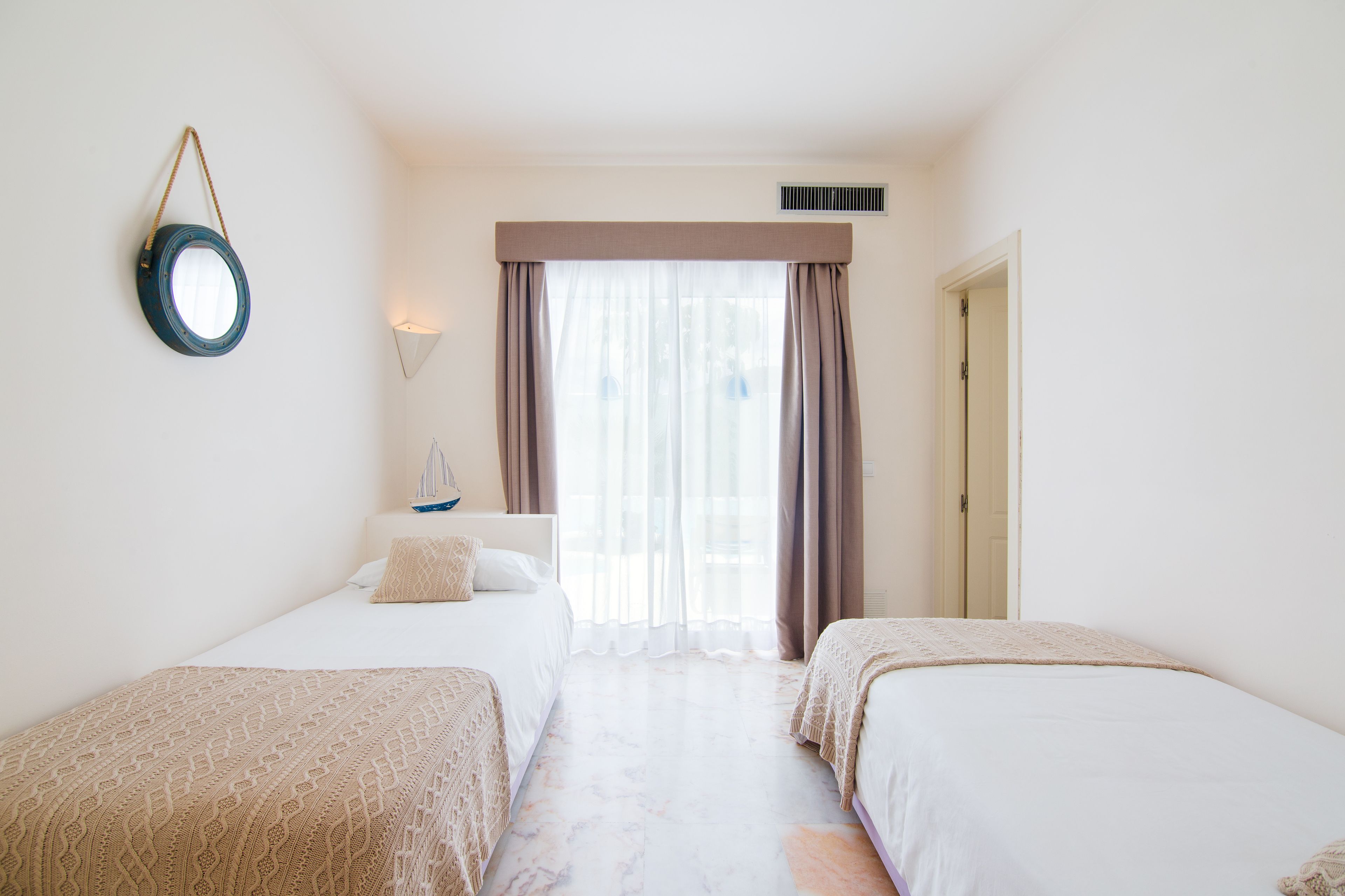 Habitación de hotel con dos camas, un espejo redondo en la pared, una ventana con cortinas y una puerta.