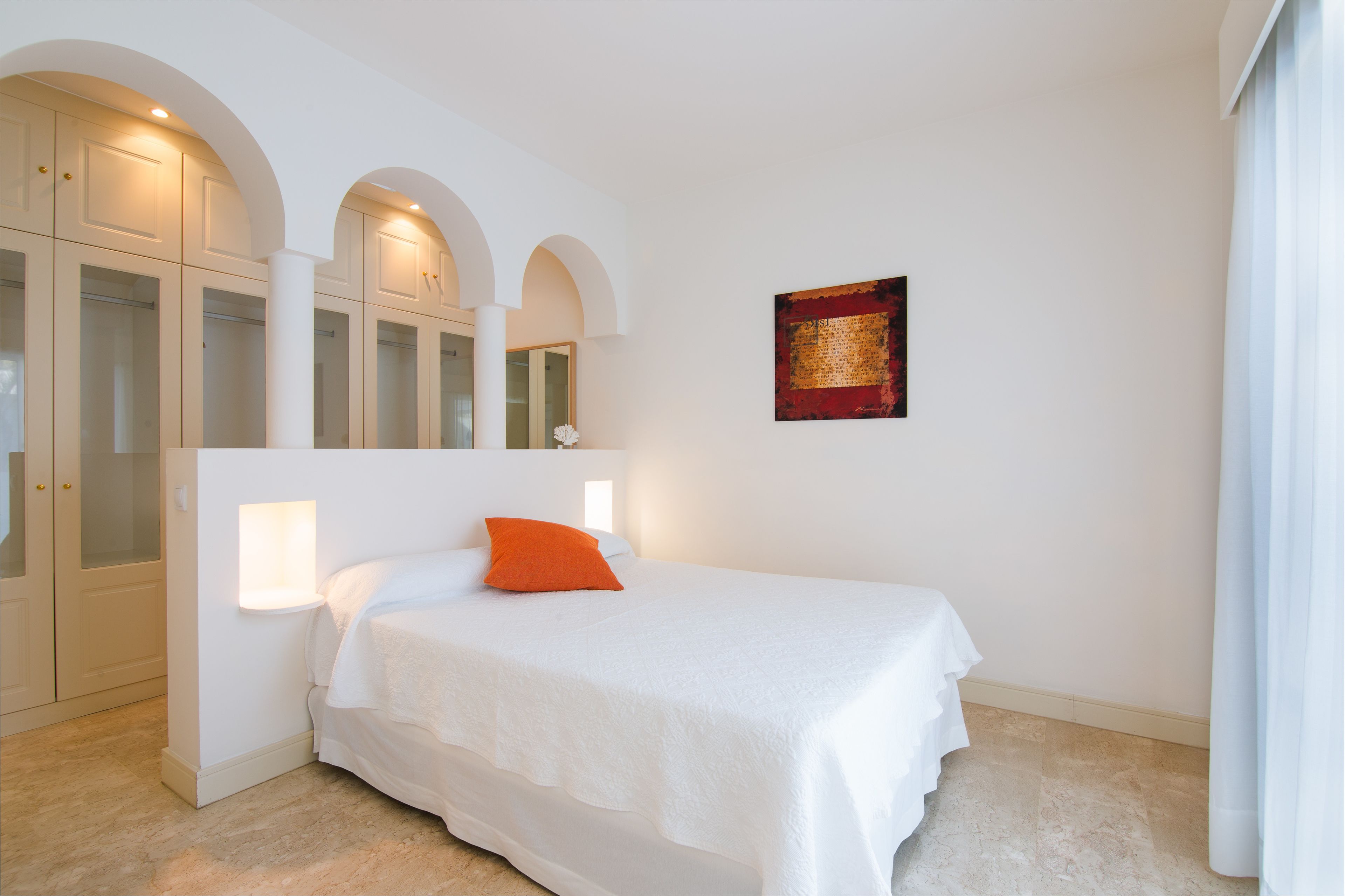 A bedroom with a bed, white walls, a red accent wall, and a window with curtains.