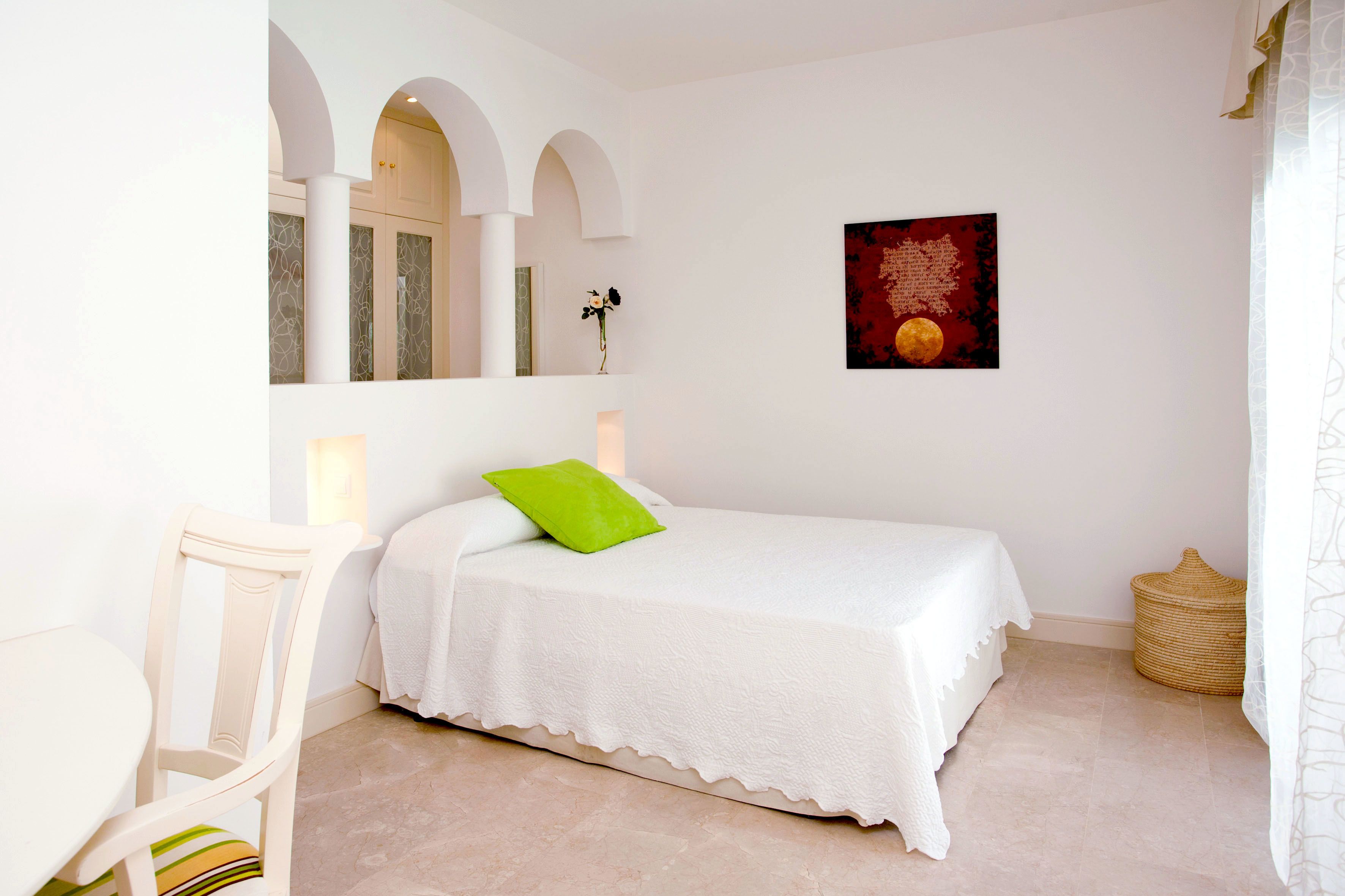 Un dormitorio con una cama, una silla, un espejo montado en la pared, un adorno mural y una cesta tejida.
