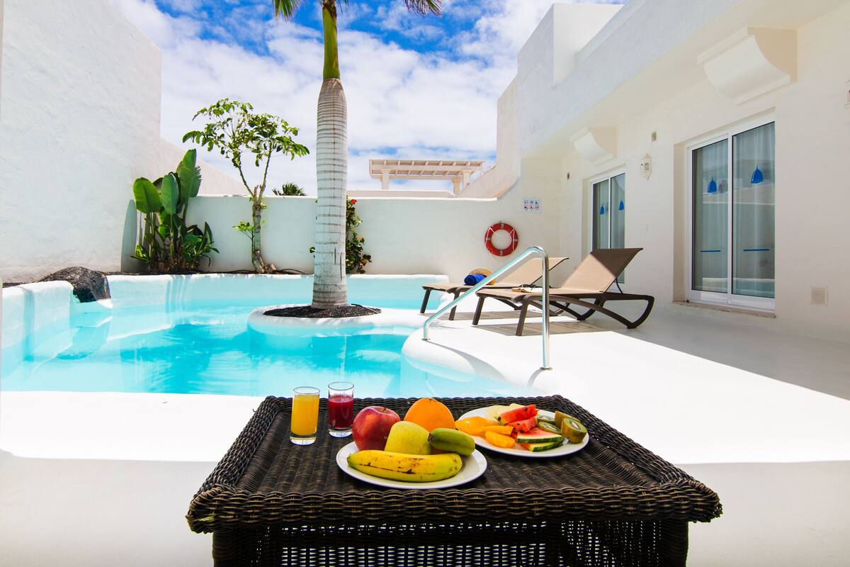 A poolside area with a table set for breakfast, including fruits and juices, and a lounge chair.