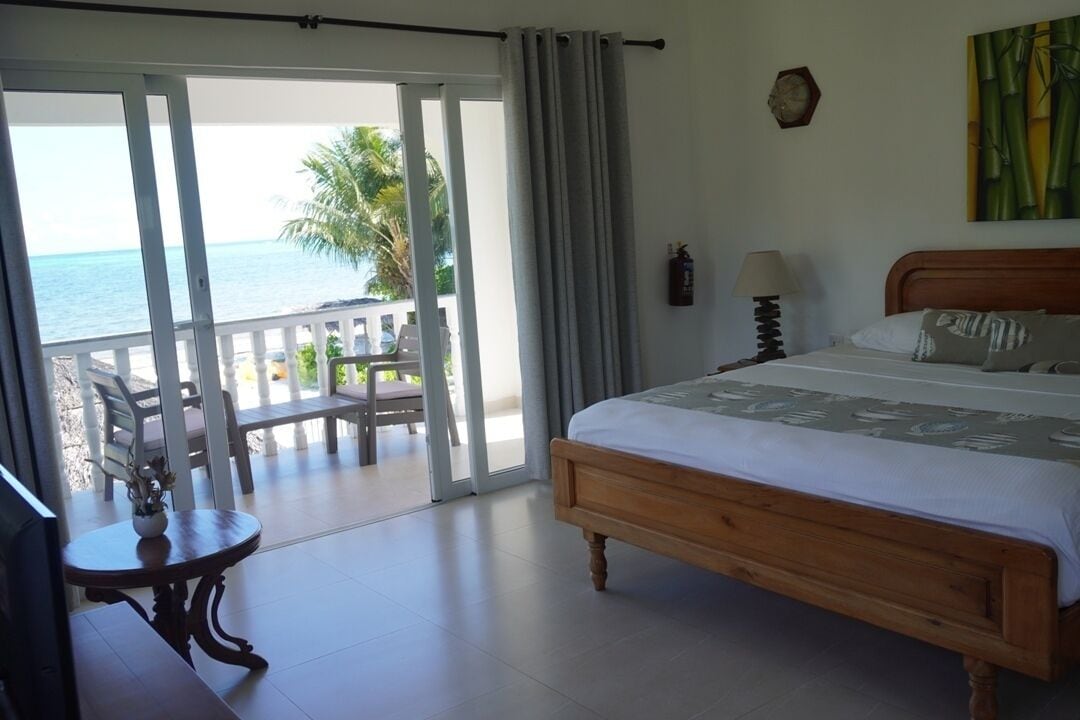 A bedroom with a wooden bed, a view of the ocean, a balcony with chairs, and a table with a vase.