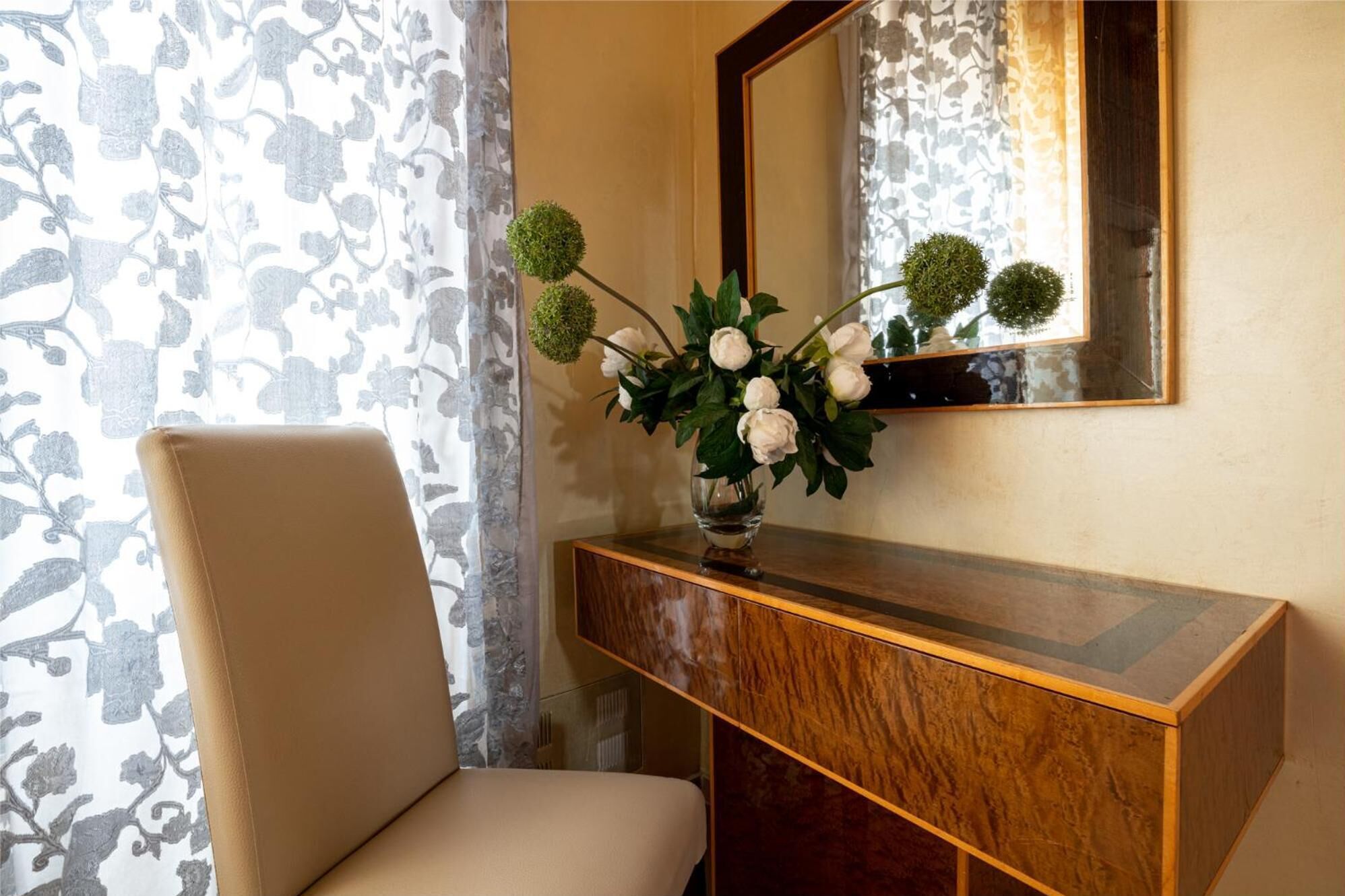 Une pièce comprenant une console en bois, un vase avec des fleurs blanches, une chaise et un miroir au mur.