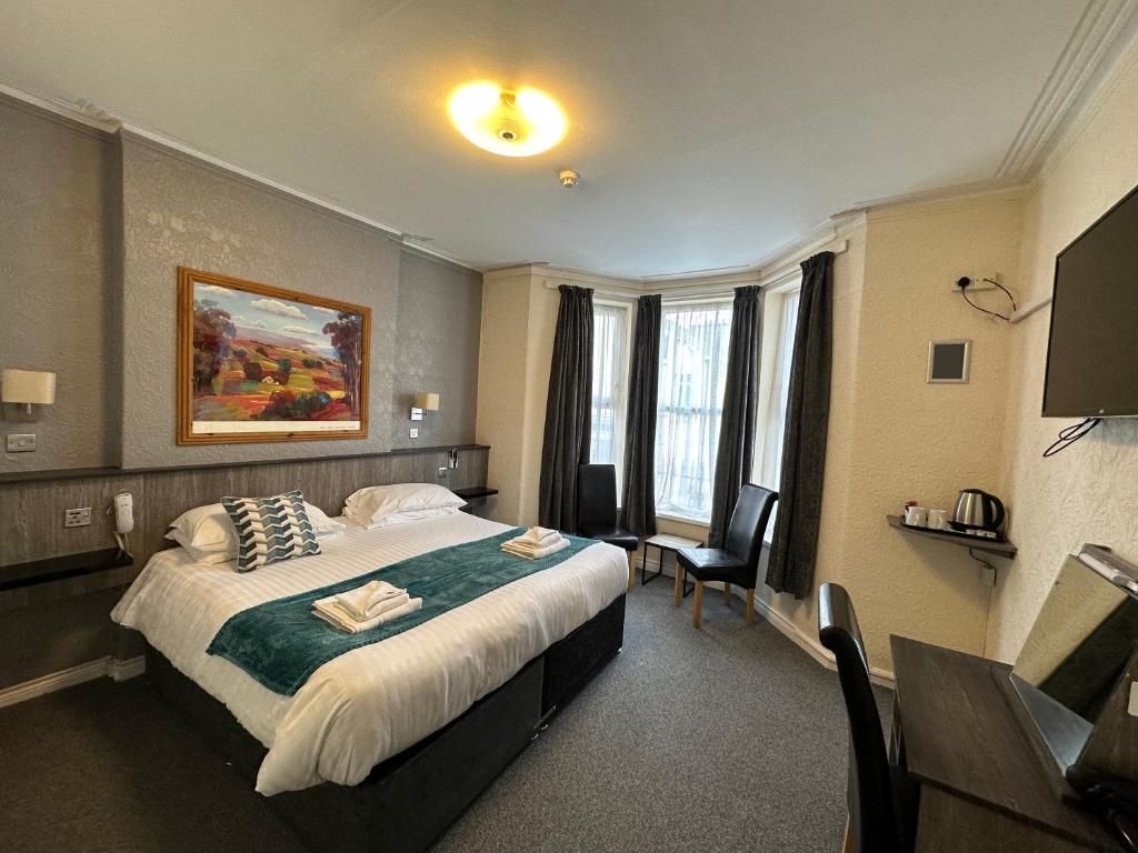 A hotel room with a large bed, a painting on the wall, a desk with a chair, and a window with curtains.
