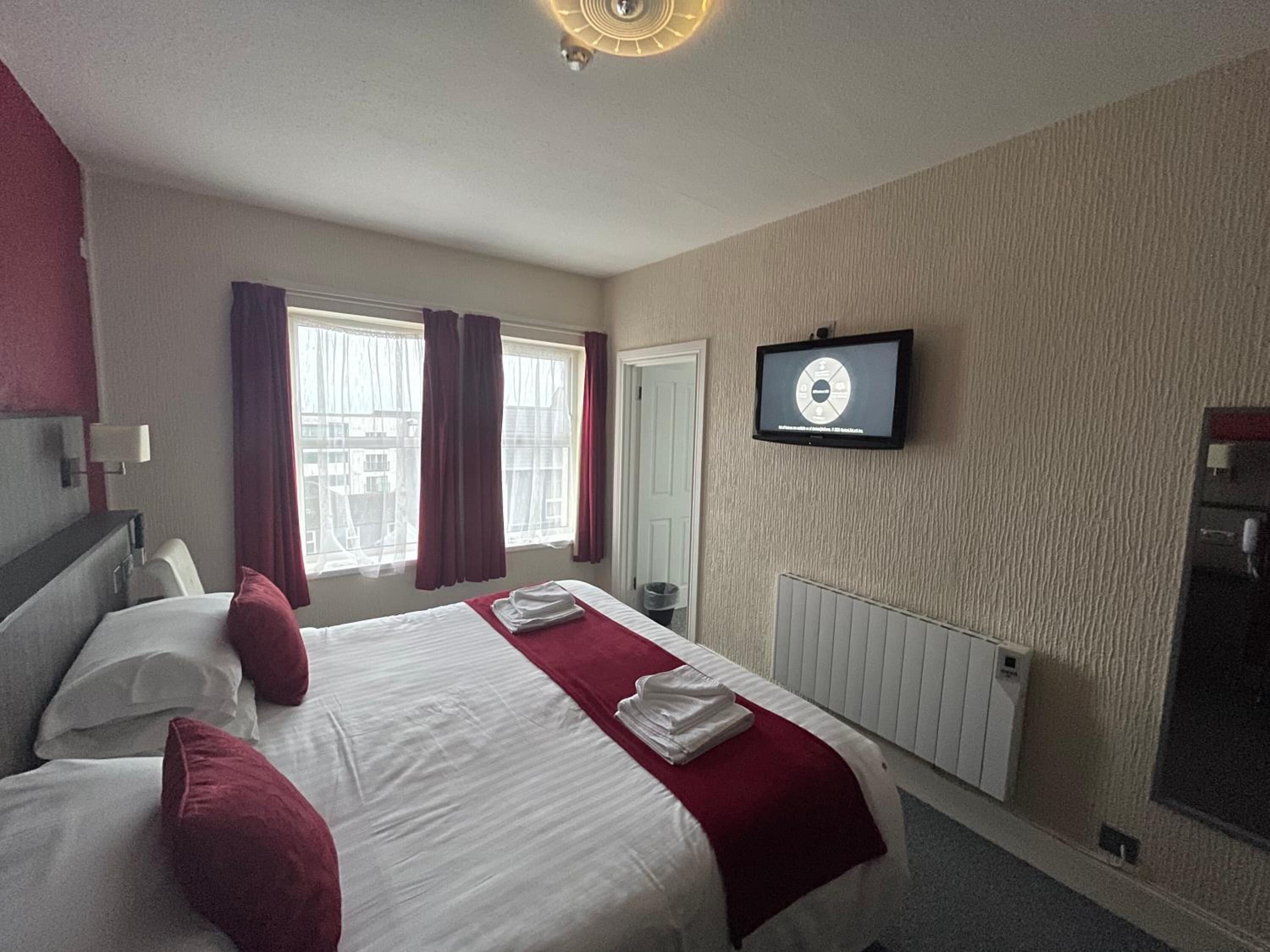 A hotel room with a bed, a TV mounted on the wall, a window with curtains, and a radiator.