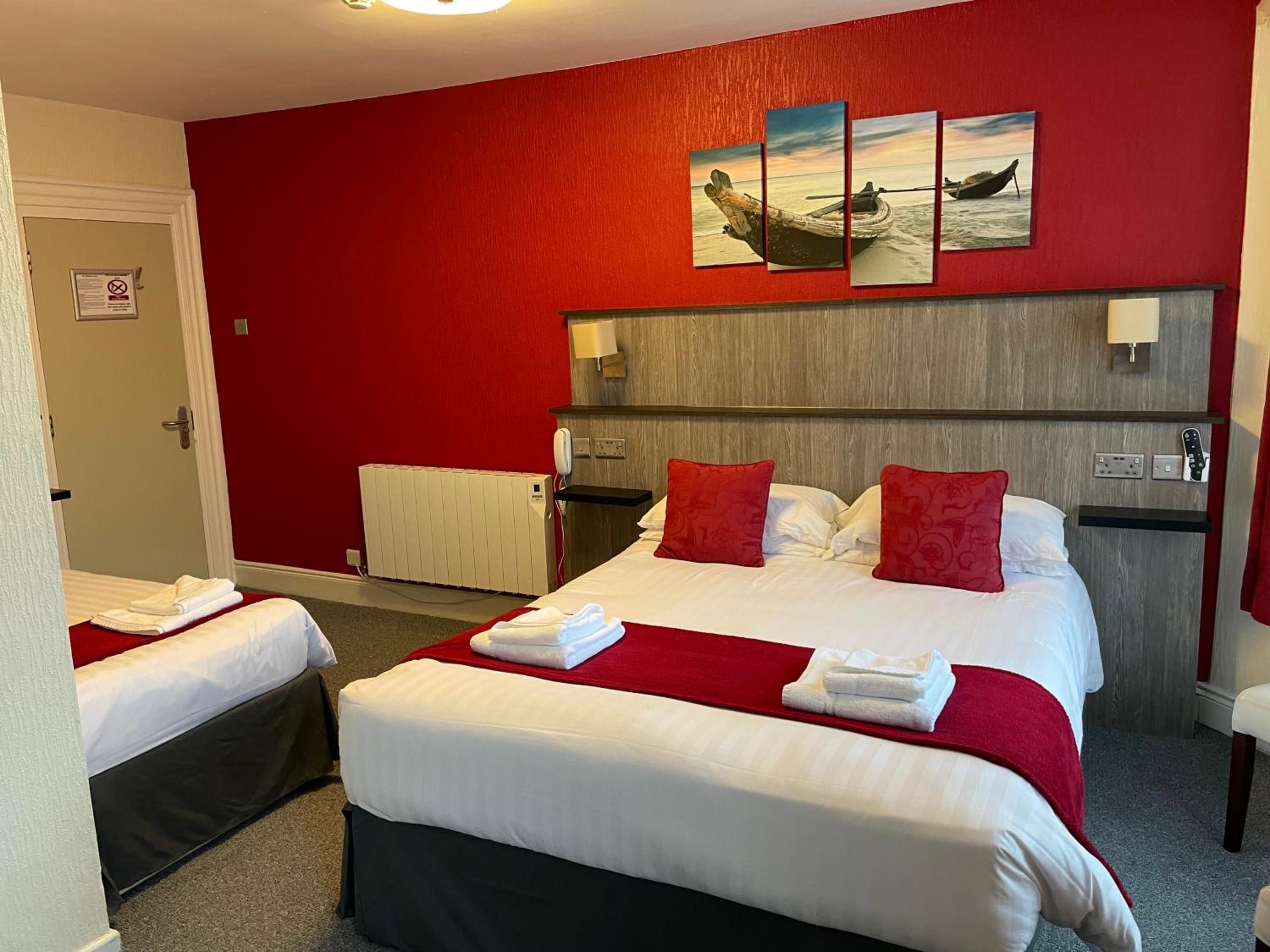 Habitació d'hotel amb una paret vermella, un llit amb roba de llit blanca i nínxols vermells, una taula de nit amb una làmpada, i un quadre d'una embarcació a la paret.