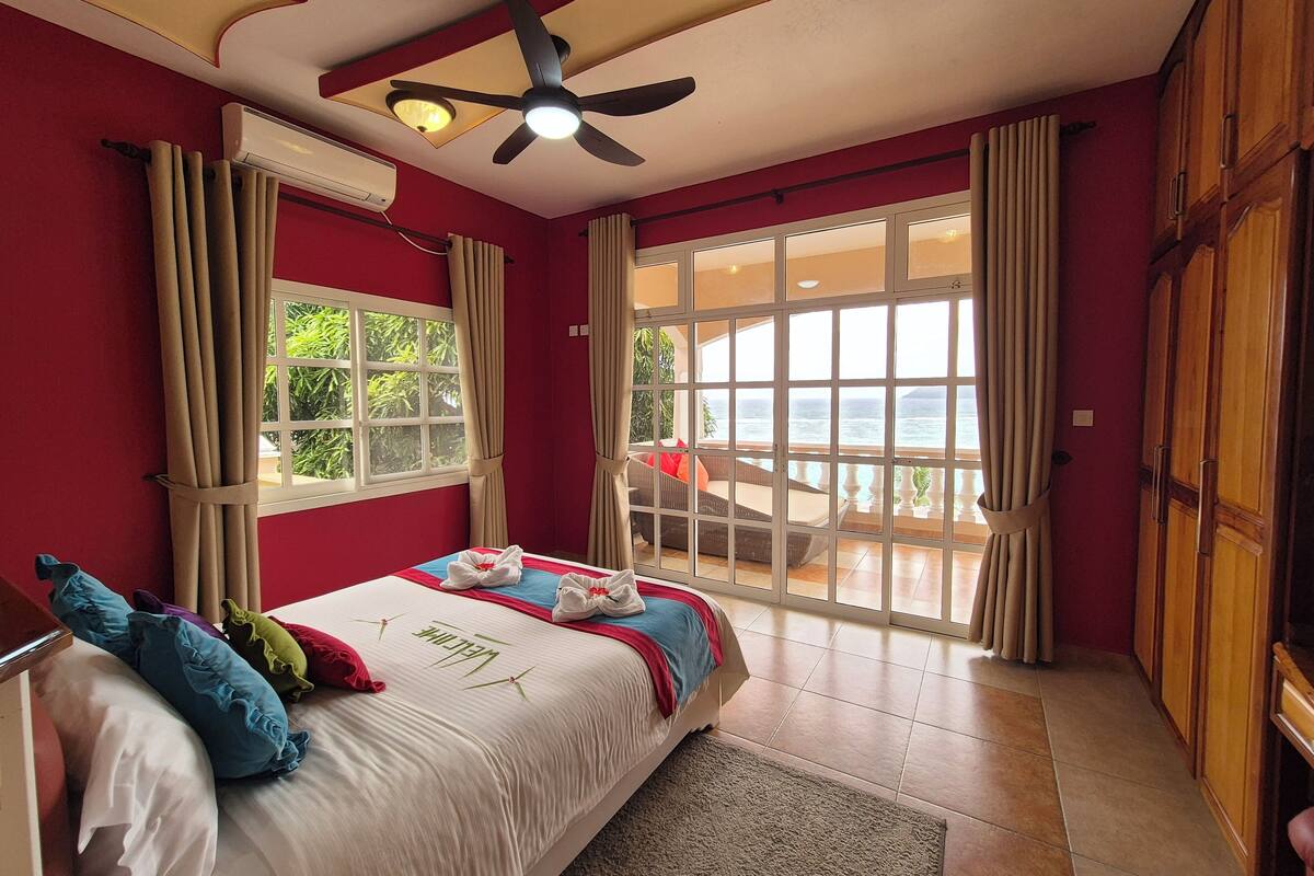 Un dormitorio con cama, ventilador de techo, ventana y vista al mar.