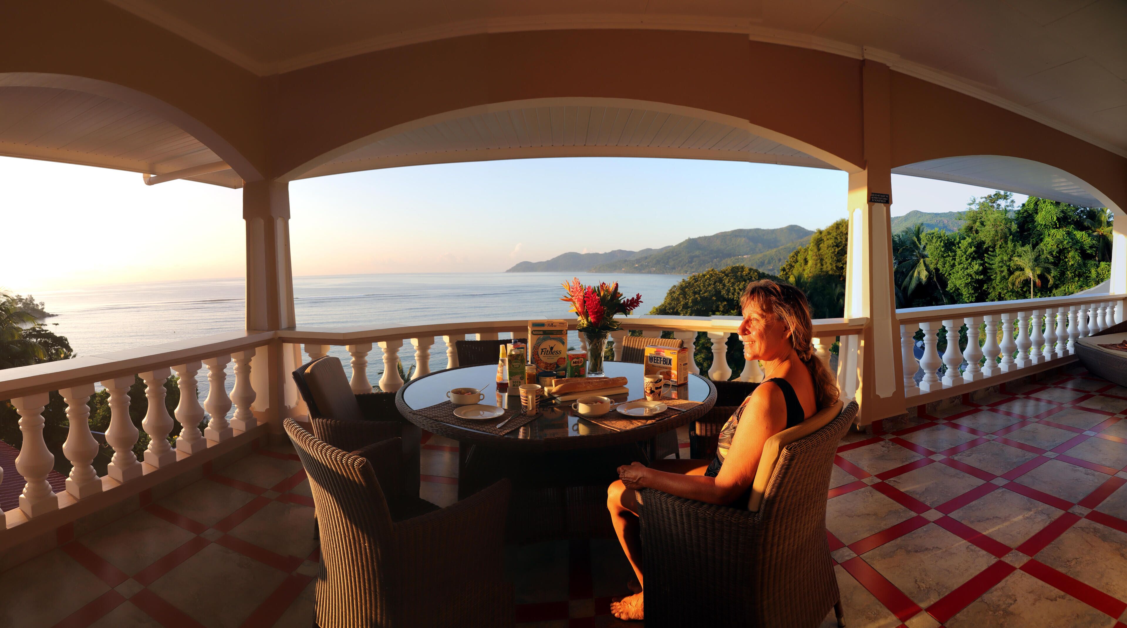 A balcony with a table set for breakfast, overlooking a scenic view of the sea and mountains.
