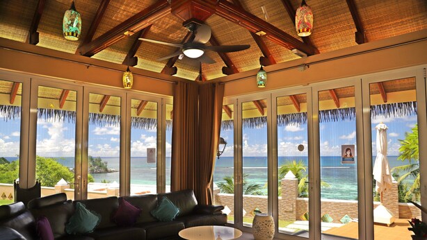 A room with a large window offering a view of the ocean, a ceiling fan, and a sofa with cushions. - Au Fond De Mer View (Mahé Island)