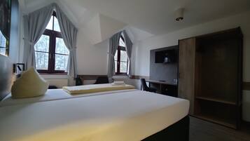 Habitación de hotel con una cama grande, dos ventanas con cortinas, un escritorio con silla, televisión y armario.